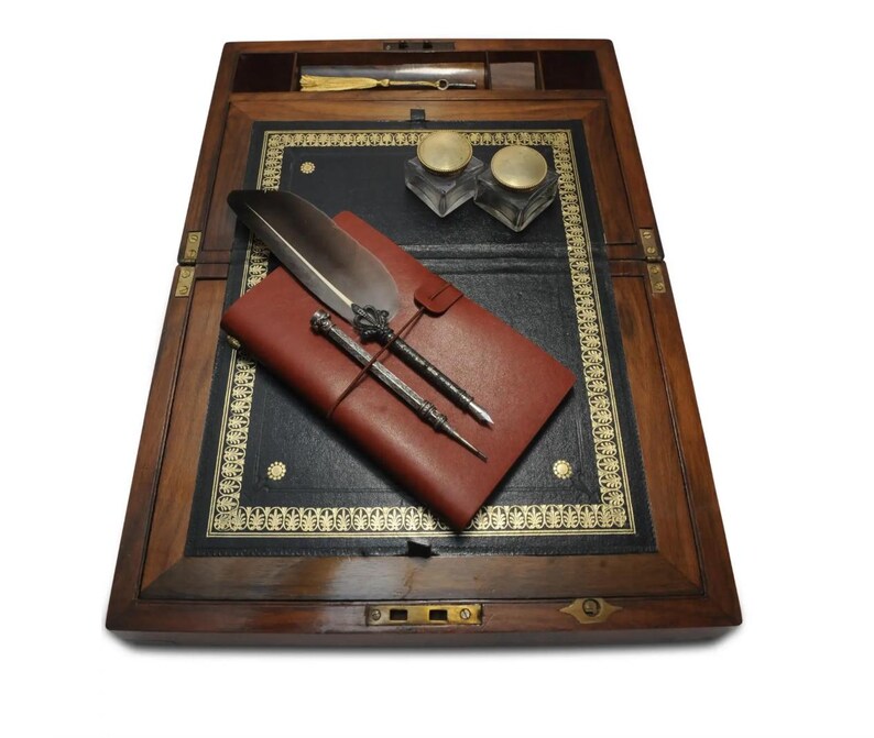 May include: A wooden writing desk with a leather writing surface. The desk is open and has a red leather journal, two inkwells, a quill pen, and two metal pens on the surface.