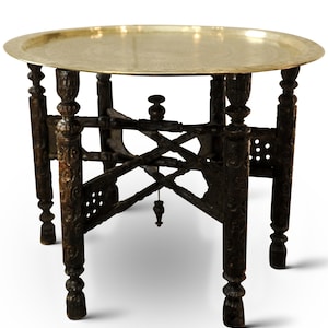 Può includere: Un tavolo decorativo in legno marrone scuro con un piano rotondo in metallo color oro. Il tavolo ha gambe intagliate in modo intricato e una base dettagliata, che mostra un'estetica vintage o antica. Il tavolo è alto circa 51 cm.