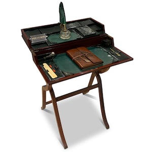 Può includere: Una scrivania da scrittura vintage con finitura in legno scuro e design a gambe pieghevoli. La scrivania presenta una superficie di scrittura in feltro verde, un diario rilegato in pelle e un calamaio di vetro con una penna verde. Ha più scomparti.