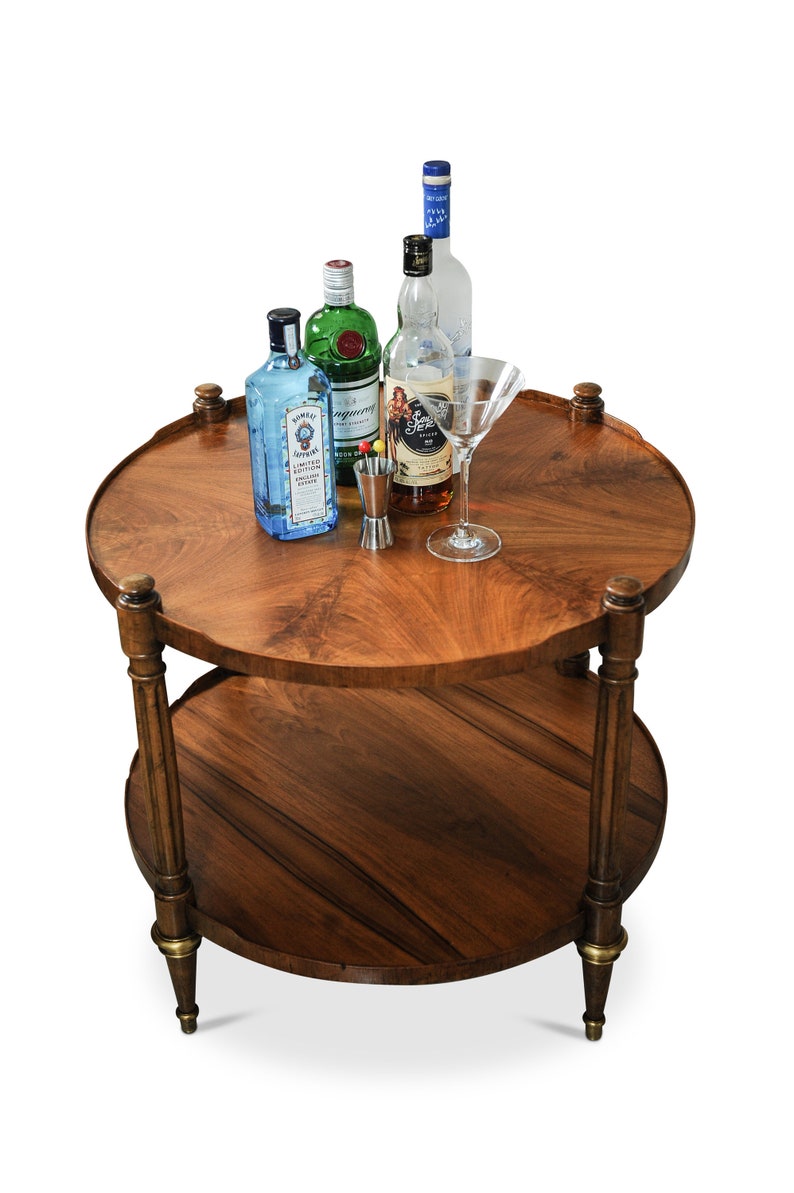 May include: A two-tiered wooden serving cart with a round top and bottom shelf. The cart has four legs and is made of dark wood with a natural wood grain. There are four bottles of liquor on the top shelf, including Bombay Sapphire gin, Tanqueray gin, Captain Morgan spiced rum, and a bottle of vodka. There is also a martini glass and a small metal jigger on the top shelf.