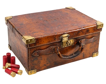 May include: A rectangular brown leather case with brass corner reinforcements, a central brass latch, and a leather handle. The name H. GREENACE is embossed on the lid. Five red shotgun shells are positioned to the left of the case.