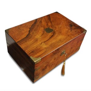 May include: A wooden writing box with a brass lock and key, a brass plate on the lid, and brass corner accents. The box is made of dark wood with a rich grain pattern.
