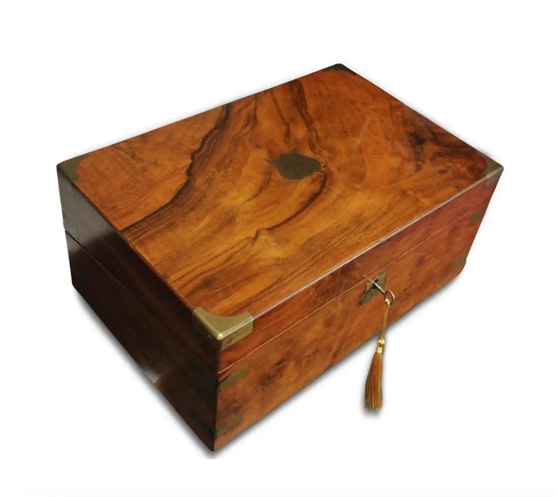 May include: A wooden writing box with a brass lock and key, a brass plate on the lid, and brass corner accents. The box is made of dark wood with a rich grain pattern.