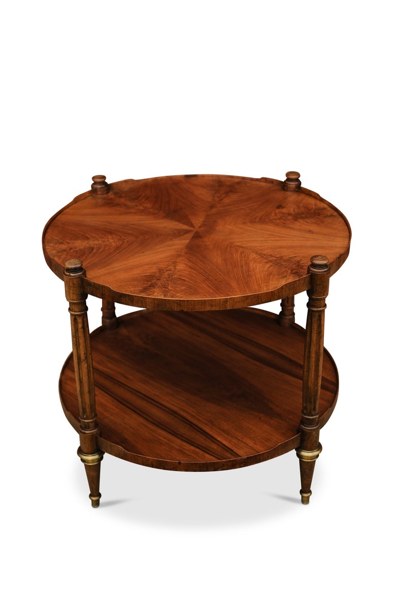 May include: A two-tiered round wooden table with a dark brown finish. The table has a scalloped edge and a decorative design on the top tier. The legs are turned and have a brass finish.