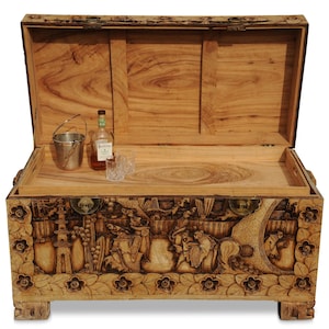 Può includere: Un forziere in legno con coperchio aperto, con intagli intricati di figure e motivi floreali. All'interno, un piccolo secchio di metallo, una bottiglia di liquore e due bicchieri sono visibili su un vassoio di legno. Il forziere è in legno chiaro.