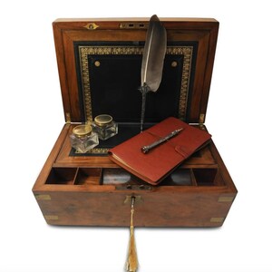 May include: An antique wooden writing box with a leather-lined interior. The box is open and contains a quill pen, a notebook, and two inkwells. The box has brass accents and a keyhole with a tassel.