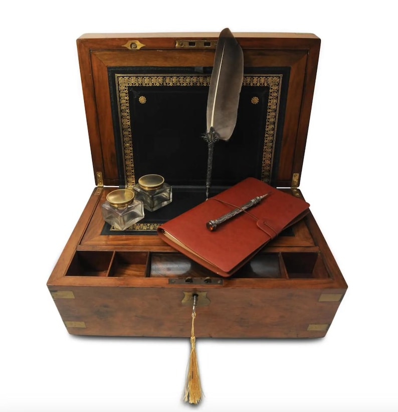 May include: An antique wooden writing box with a leather-lined interior. The box is open and contains a quill pen, a notebook, and two inkwells. The box has brass accents and a keyhole with a tassel.