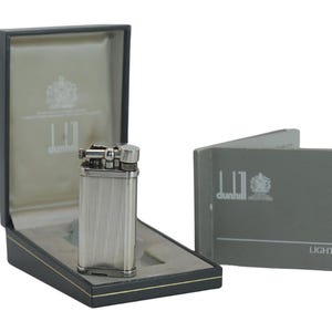 May include: A silver-coloured, vertically ribbed lighter with a flip-top design, presented in a black case with a grey interior. A matching grey booklet with the word "Lighters" is also included.