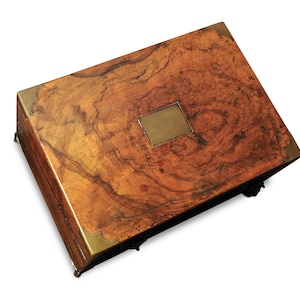 Puede incluir: Una caja de madera rectangular con tapa con bisagras. La caja está hecha de una madera de tonos cálidos con un patrón de vetas arremolinadas. Tiene esquinas de latón y una placa de latón en la tapa. La caja está elevada sobre pequeños pies.