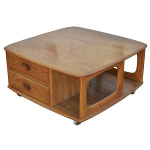 Op de afbeelding: Een salontafel in mid-century stijl van lichtbruin hout. Het heeft twee lades aan één kant en een open plank aan de andere kant. De tafel staat op kleine wieltjes. Afmetingen zijn ongeveer 90 cm x 90 cm.