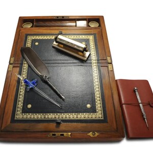 May include: A wooden writing desk with a leather writing surface, a quill pen, a glass dip pen, and a brass inkwell. The desk has a hinged lid and a brass lock.