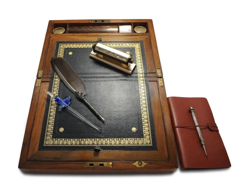 May include: A wooden writing desk with a leather writing surface, a quill pen, a glass dip pen, and a brass inkwell. The desk has a hinged lid and a brass lock.