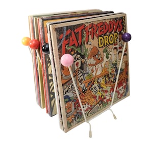 Pode incluir: Um suporte para discos de metal branco com cinco botões redondos coloridos segura uma pilha de discos de vinil. O disco superior tem uma ilustração de desenho animado de um homem com bigode e as palavras "Fat Freddy's Drop".