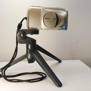 May include: A silver Olympus digital camera mounted on a black tripod. The camera has a retractable lens and the brand name is visible. The tripod has three legs and a black cord. The camera is designed for photography.