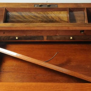 May include: An antique wooden writing desk with a hinged top, revealing a compartment with two small drawers and a larger open space. The desk has a rich brown finish and intricate wood grain.