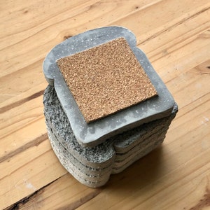 May include: A stack of gray concrete coasters shaped like slices of bread. The top coaster has a cork insert.