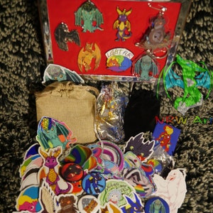 May include: A collection of dragon-themed pins, stickers, and buttons. The items are arranged on a brown surface and include a variety of colors, including green, blue, purple, and red. The text "LGBT FUR" is visible on one of the pins.