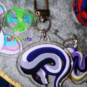 May include: A purple, white, and black snake charm with a silver key chain and a silver clasp. The charm is made of clear acrylic and has a glossy finish.
