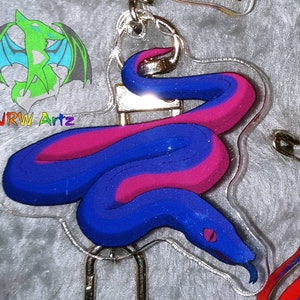 May include: A blue and pink snake charm with a black eye, hanging from a silver key chain. The charm is made of clear acrylic and has a glossy finish.
