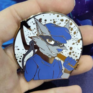 Puede incluir: Un pin de esmalte de color dorado con un lobo de dibujos animados que lleva una gorra azul y una camisa azul. El lobo sostiene un bate de madera y tiene una expresión traviesa en su rostro. El fondo es blanco con puntos dorados.