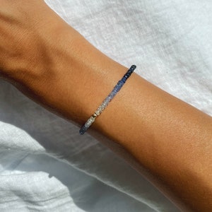 May include: A delicate bracelet made of small, graduated blue gemstones. The bracelet is strung on a thin gold chain and features a subtle ombre effect, transitioning from light to dark blue.