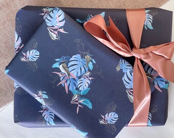 Puede incluir: Un papel de regalo azul marino con un estampado tropical con flores azules y rosas, hojas verdes y pájaros. El papel de regalo está atado con una cinta de satén de color cobre.