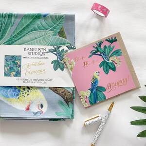May include: A blue tea towel featuring a tropical bird and leaf design. The towel is packaged with a pink card with a similar design and the text "Hi x Hi" and "Hooray!" in gold lettering.