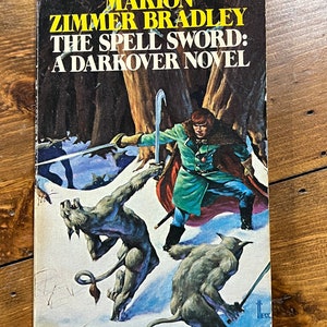 May include: A book cover featuring a fantasy illustration of a woman in green and red clothing holding a sword, facing two wolf-like creatures in a snowy forest. The book title is "The Spell Sword: A Darkover Novel" by Marion Zimmer Bradley.