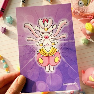 May include: A rectangular art print depicting a stylized character with a white head, pink body, and yellow accents against a purple gradient background. The print is held by a hand, with the artist's handle, @pinkeepokie, visible. Crayons and decorative items surround the print.