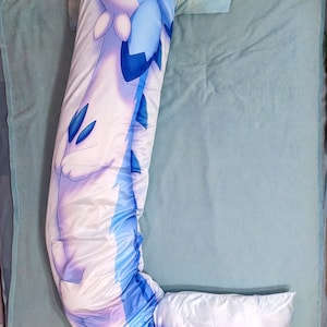 K&ouml;nnte beinhalten: Ein langes, wei&szlig;es und blaues Kissen mit einem Cartoon-Drachen-Design. Der Drache ist in einer Schlafposition zusammengerollt, mit seinen Fl&uuml;geln um seinen K&ouml;rper gewickelt. Das Kissen liegt auf einem blauen Bettbezug.