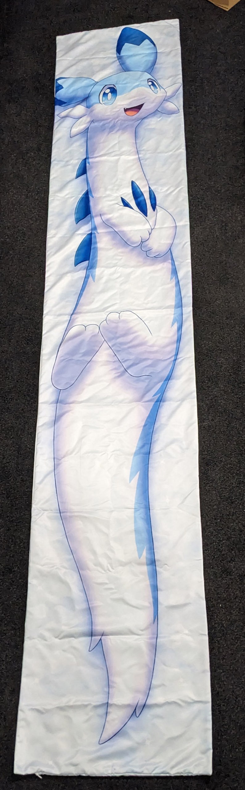 K&ouml;nnte beinhalten: Ein langes wei&szlig;-blaues Kissen mit einem Cartoon-Bild eines blau-wei&szlig;en Drachen mit einem l&auml;chelnden Gesicht.