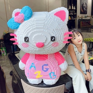 May include: A large white Hello Kitty figurine made of plastic building blocks. The figurine is wearing a pink dress with colourful letters and a blue bow on its head. A young girl is sitting next to the figurine.
