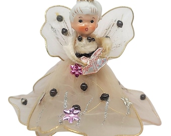 May include: A figurine of a small angel with white hair, wearing a beige tulle dress adorned with black beads, silver glitter accents, and pink flower embellishments. The angel has translucent wings and holds a small book with the text Noel.