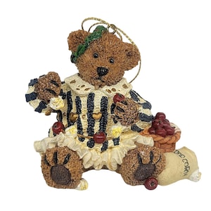 May include: A brown teddy bear figurine wearing a striped outfit with a ruffled collar and holding a basket of red apples. The bear has a green wreath on its head and a small sack of corn.