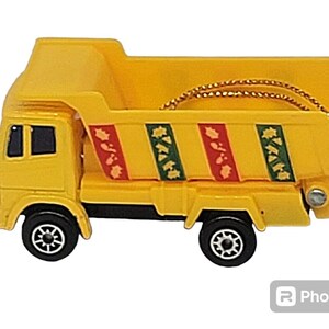 May include: A yellow toy dump truck with a red and green patterned bed. The truck has a gold string attached to it.