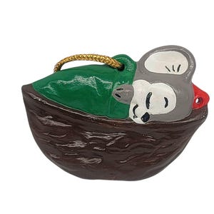 May include: A ceramic Christmas ornament shaped like a mouse sleeping in a brown nut shell. The mouse is gray with a white belly and a red nose. The nut shell is brown and has a green leaf on top. The ornament has a gold string for hanging.
