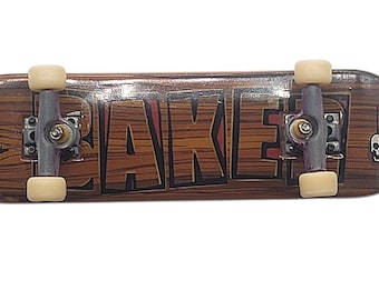Könnte beinhalten: Ein Mini-Fingerboard mit Holzmaserung. Das Wort "LAKE" ist in großen, braunen Buchstaben mit roter Umrandung gedruckt. Das Board hat silberne Achsen und beige Rollen. Eine kleine Totenkopf-Grafik befindet sich auf der rechten Seite.