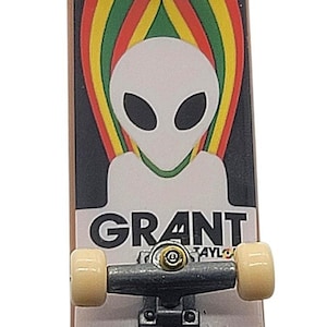 May include: A miniature skateboard with a black top featuring an alien graphic and the name "GRANT". The design includes a flame-like pattern with red, yellow, and green stripes. The wheels are beige, and the board has the text "CARRY THE TORCH".