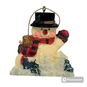 May include: A white snowman Christmas ornament with a black top hat, red and black scarf, and a red mitten. The snowman is sitting in a snowdrift with green pine branches.