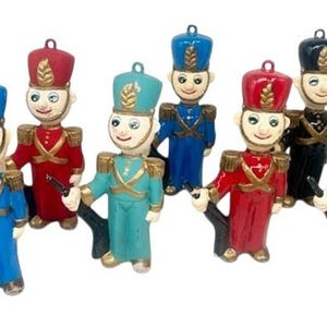May include: A collection of colorful toy soldier figurines. Each soldier is painted with a different color uniform, including green, blue, red, and black. They are holding black rifles and have gold accents on their uniforms.