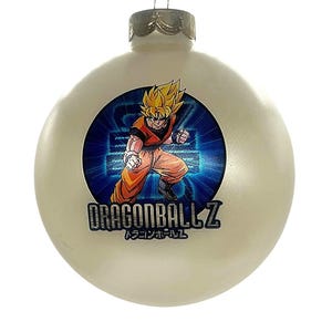 May include: A cream-colored Christmas ornament featuring a Dragon Ball Z graphic. The graphic depicts a character in an orange and red outfit with spiky yellow hair, set against a blue background. The text "DRAGONBALL Z" is displayed below the character.