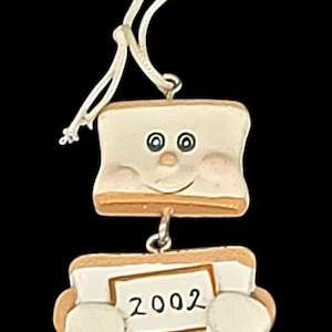 May include: A ceramic ornament shaped like a smiling s'more, with a white marshmallow face and a graham cracker base. The bottom section displays the year "2002". It has a white string for hanging, perfect for holiday decor.