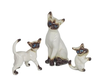 May include: Three ceramic Siamese cat figurines. The cats have white bodies, brown faces, ears, and paws, and blue eyes. One cat is sitting, one is standing, and one is in a playful pose.