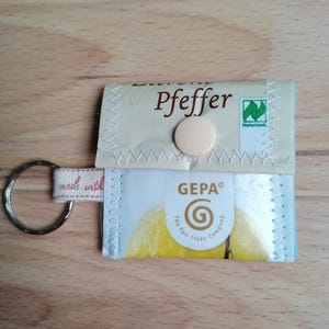 Könnte beinhalten: Ein kleines, handgefertigtes Schlüsselanhänger-Portemonnaie mit einem silbernen Schlüsselring. Das Portemonnaie besteht aus recycelten Verpackungen mit dem Text "Pfeffer" und "GEPA" mit einem Knopfverschluss. Das Etikett lautet "made with".