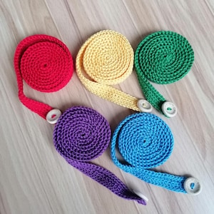 Pode incluir: Cinco suportes de máscara de crochê coloridos em vermelho, amarelo, verde, roxo e azul. Cada suporte é enrolado e tem uma alça com fecho de botão. Os botões brancos têm um pequeno desenho.