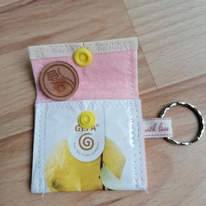 Könnte beinhalten: Eine kleine, offene rosa Filztasche mit gelbem Druckknopfverschluss, die eine Holzmünze und ein weißes Päckchen mit einem Zitronenmotiv enthält. Ein Metall-Schlüsselring ist an einem Stoffetikett mit der Aufschrift "with love" befestigt.