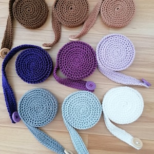 Pode incluir: Uma coleção de tiras de máscara de crochê em várias cores, incluindo marrom, roxo e azul. Cada tira é enrolada e possui um fecho de botão. As tiras são projetadas para serem usadas ao redor da cabeça.
