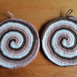 Op de afbeelding: Twee gehaakte onderzetters in bruin, wit en roze met een spiraalpatroon.