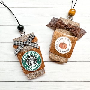 May include: Two gold glitter coffee cup charms with black and white checkered ribbon bows. One charm features the Starbucks logo and the text "Starbucks Coffee". The other charm features a pumpkin and the text "Autumn is calling, leaves are falling".