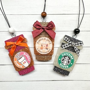 May include: Three coffee cup-shaped car air fresheners with glitter and different designs. The first is pink with orange ribbon and the text "Dunkie Junkie". The second is gold with a brown ribbon and the text "Pumpkin Spice & Everything Nice". The third is black with a white and black ribbon and the text "Caffeine Queen".